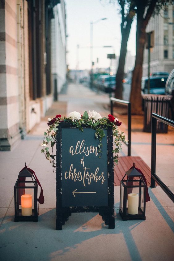 a small and pretty wedding sign decorated with greenery and blooms and black vintage lanterns on both sides is lovely