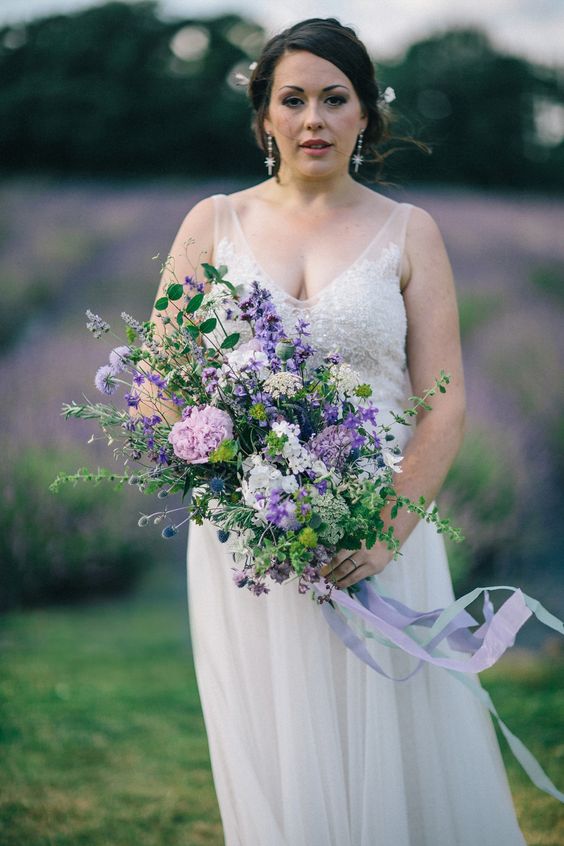 a textural and romantic purple wedding bouquet of purple, lilac, pink and neutral blooms, lots of greenery and thistles and long ribbon