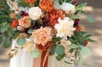 a bold fall wedding bouquet that includes orange, blush and neutral blooms, greenery, gilded leaves and berries plus long ribbons