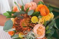 a bold wedding bouquet with orange and yellow ranunculus, a pincushion protea, coral peony roses, billy balls, greenery and air plants