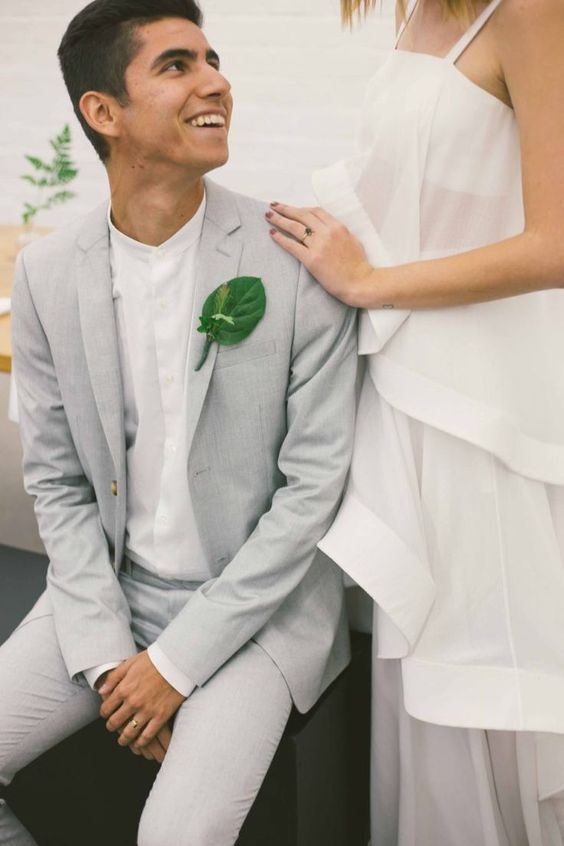 a chic minimalist groom's outfit wiht a dove grey pantsuit, a white shirt and a green leaf boutonniere for a tropical wedding