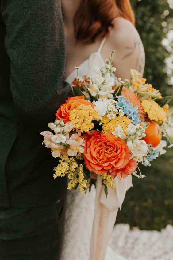 a colorful fall wedding bouquet with yellow blooms, white, blue and blush ones, an apple and mimosas plus long ribbon
