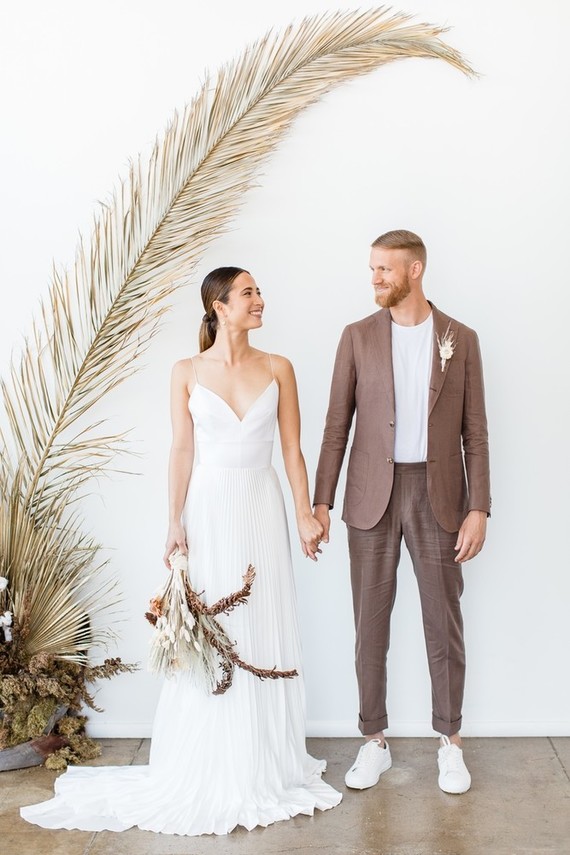 a taupe minimalist pantsuit of linen, a white t-shirt and white sneakers plus a dried flower boutonniere are a great combo for a tropical wedding
