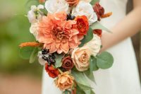 an exquisite wedding bouquet with white and orange roses, a large orange dahlias, berries and greenery with an eye-catchy shape