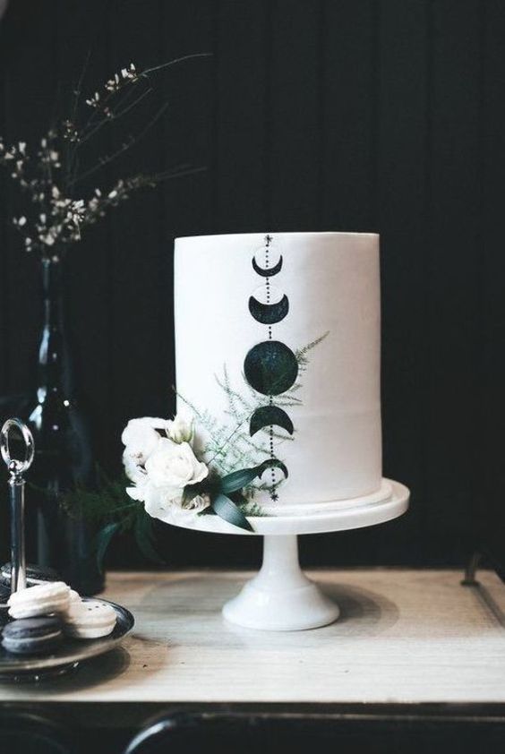 a white wedding cake with black moon phases and white blooms and greenery is a chic idea for a boho celestial wedding
