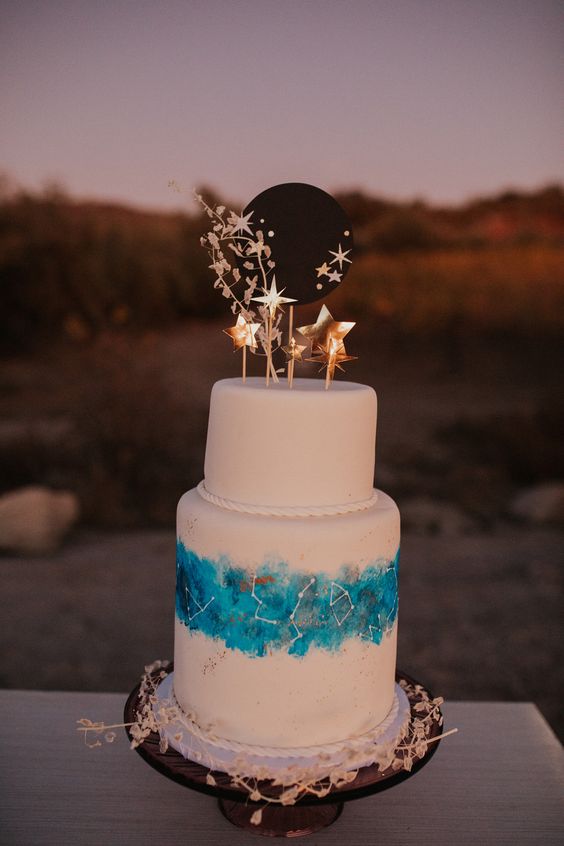 a white wedding cake decorated with turquoise paint and white constellations, with gold stars and a black moon on top