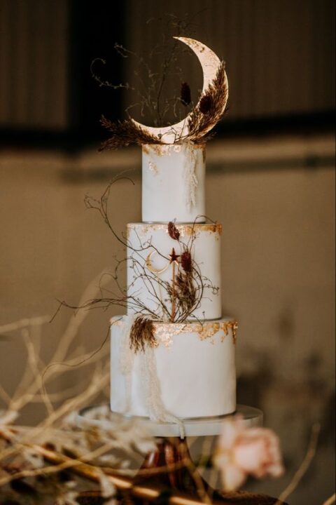a white wedding cake with gold leaf, a half moon topper, dried grasses is a lovely idea for a neutral celestial wedding