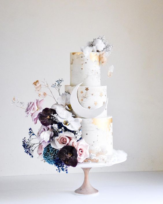 a white wedding cake with gold stars and gold leaf, constellations, a half moon, with dark and pastel blooms and cotton on top
