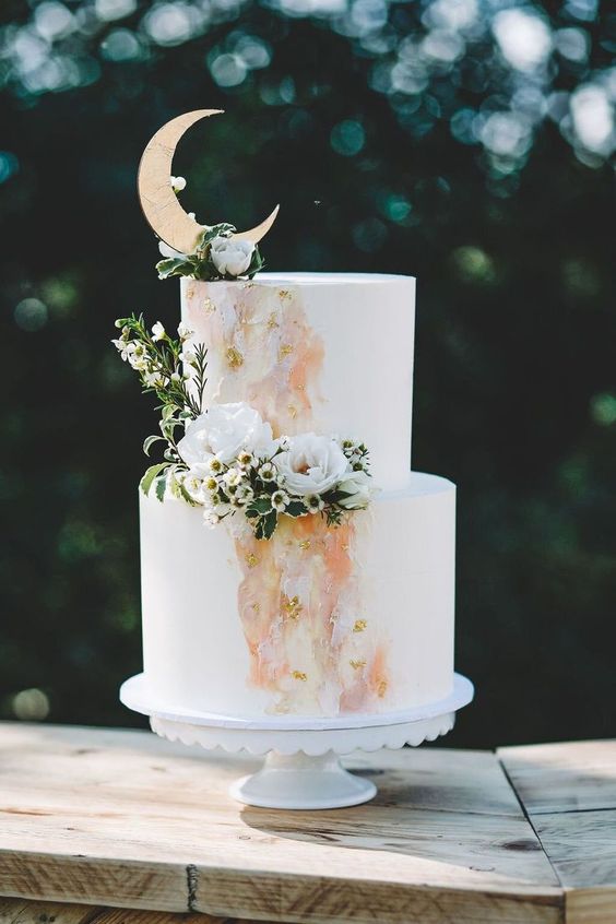 a white wedding cake with peachy, yellow and blush watercolors, white blooms and greenery and a half moon cake topper