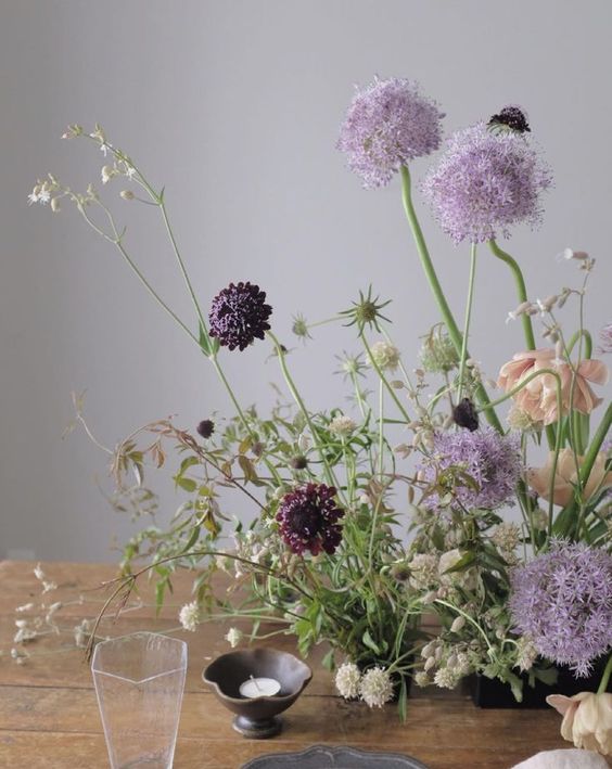 a refined and dimensional wedding centerpiece with lots of greenery, allium, deep purple blooms and thistles is beautiful