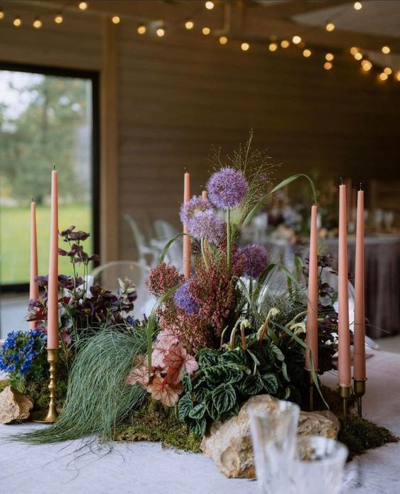 a unique secret garden wedding centerpiece of greenery and foliage, allium and bold blue flowers and pink candles