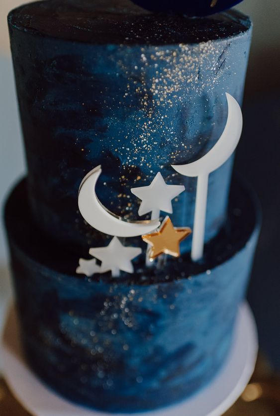 a navy wedding cake decorated with gold sparkles, gold and white stars and moon cake toppers placed on one side for a celestial wedding