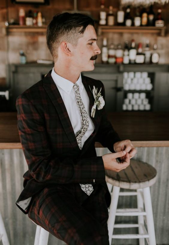 a burgundy and black plaid pantsuit, a white shirt, a neutral floral tie and a white floral boutonniere are a chic and contrasting look