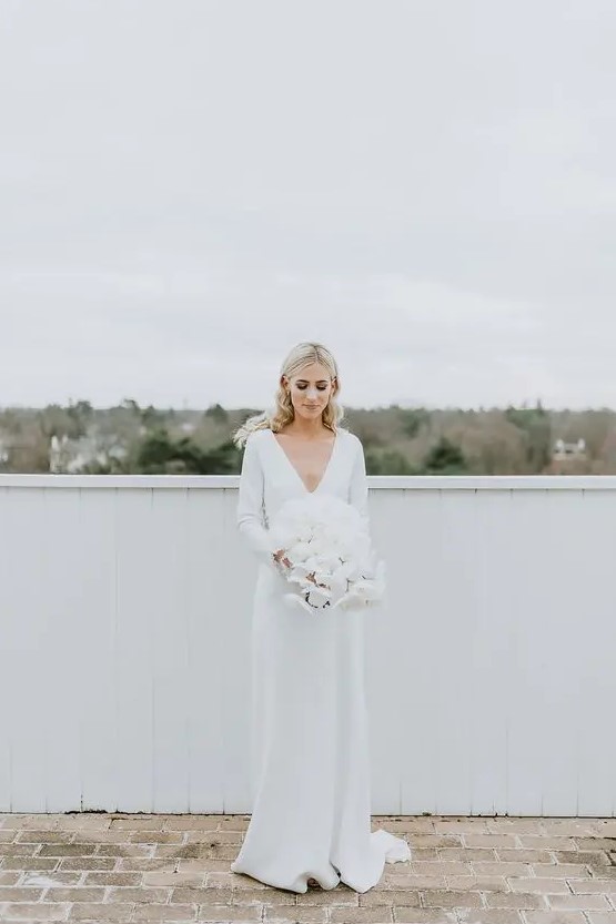 a pretty and simple minimalist winter bridal look with a plain wedding dress with a deep V-neckline, long sleeves and a train plus a lush white wedding bouquet