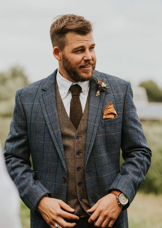 a stylish winter groom's look with a white shirt, a brown plaid waistcoat, a blue plaid blazer, a rust-colored handkerchief and a boutonniere