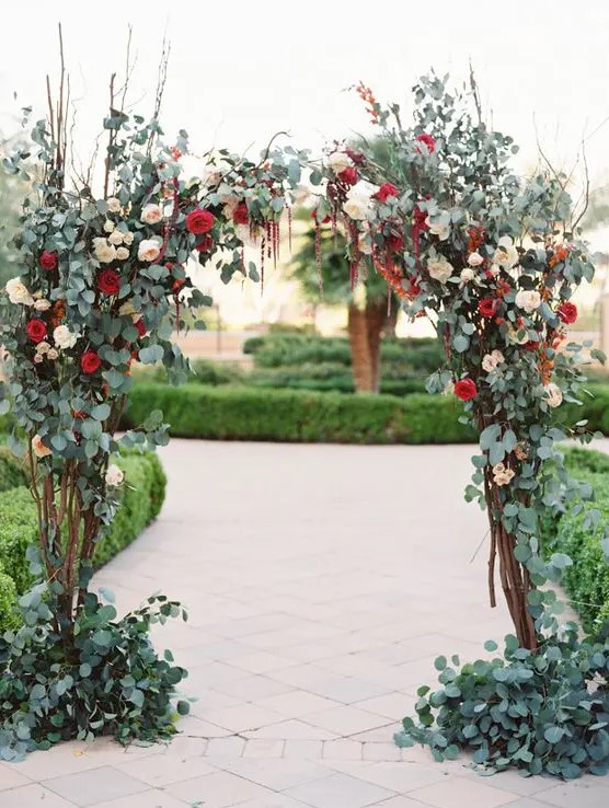 a chic fall wedding arch covered with greenery, blush and red blooms plus some twigs for a more eye catchy looks is a very cool idea for an autumen garden wedding