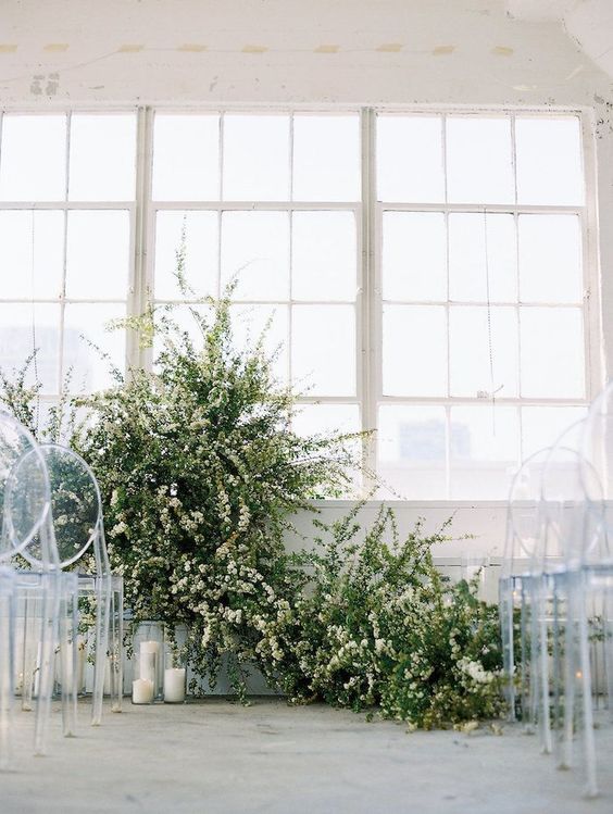 a minimalist winter wedding ceremony space done with greenery and white blooms, pillar candles and clear chairs
