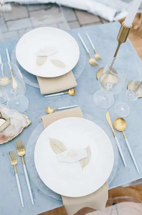a beautiful winter wedding tablescape with a blue tablecloth, clear chargers and white porcelain, gold candleholders and cutlery and tan napkins