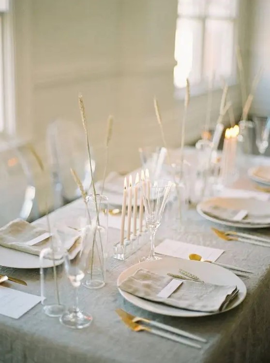 a delicate minimalist wedding tablescape with a grey tablecloth and napkins, white chargers and cutlery, some grasses in vases and tall and thin candles