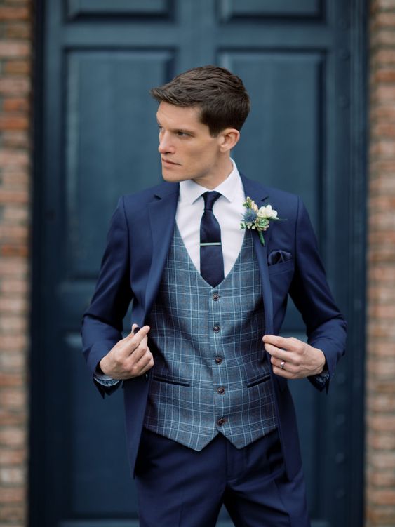 a navy pantsuit, a grey plaid waistcoat, a white shirt, a navy tie and a white boutonniere are a timelessly stylish and chic look for a groom