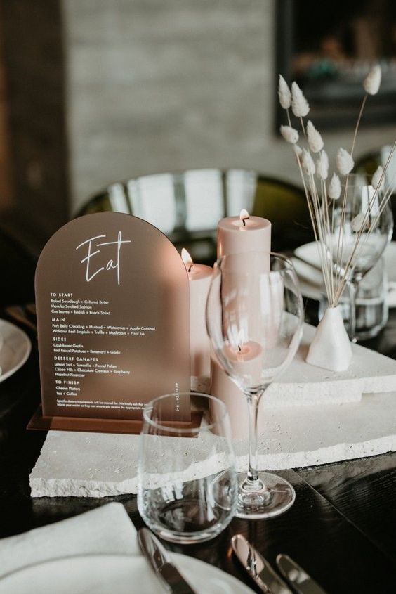 a minimalist winter wedding table with a copper arched menu, dried grasses, white stone pieces stacked and blush candles