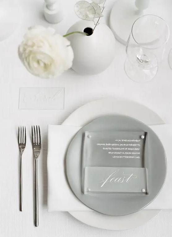 a neutral minimalist wedding table setting with white linens, a grey plate, white blooms in a round vase and simple cutlery