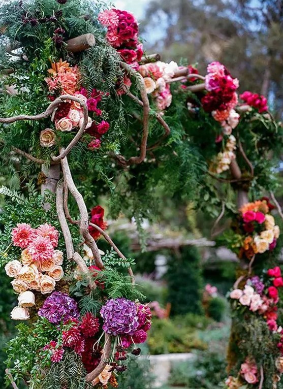 a secret garden wedding arch decorated with greenery and evergreens, bold blooms in hot pink, purple, burgundy, blush blooms and branches weaving the arch