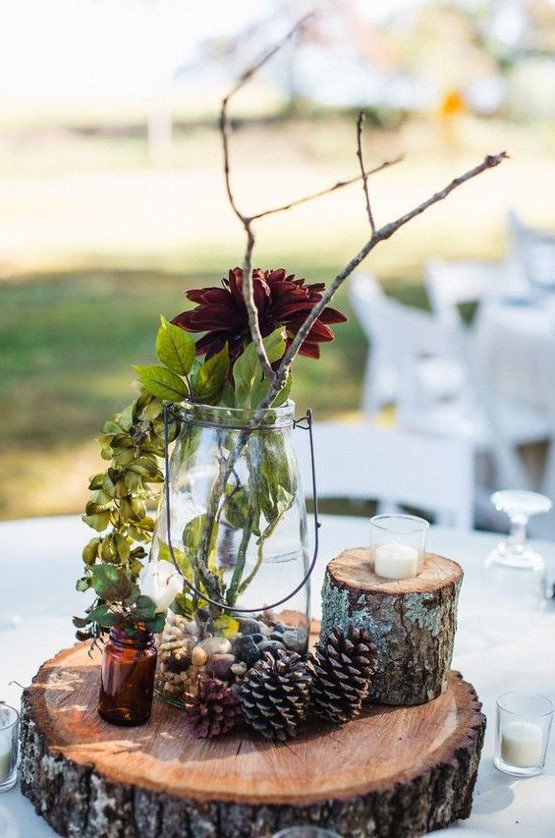 a fall wedding centerpiece with a wood slice, pinecones, greenery, branches and burgundy blooms looks very cozy and rustic