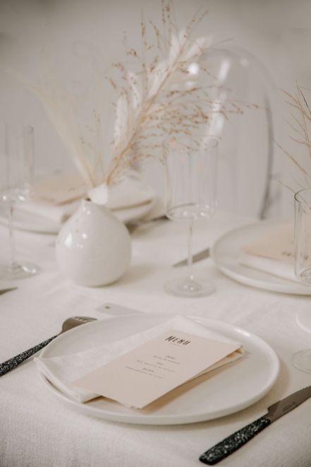 a neutral winter wedding table with white porcelain, dried grasses in vases, cutlery with black handles and neutral menus