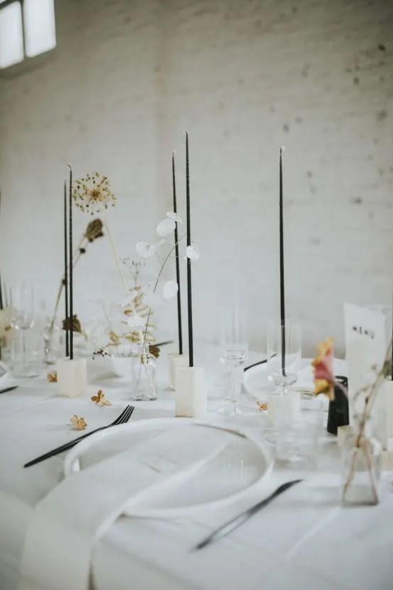 a stylish minimalist wedding tablescape with white plates and linens, white candleholders, tall and thin black candles and dried and fresh blooms