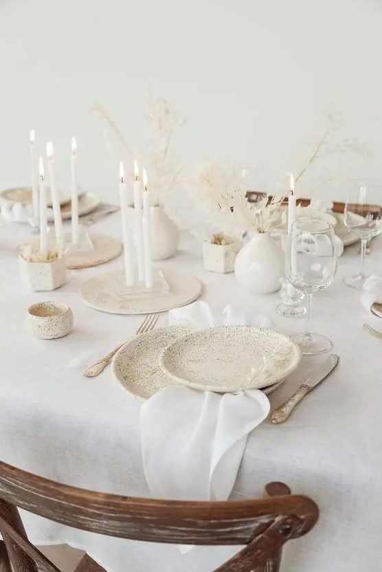an airy white winter wedding tablescape with speckled plates, tall and thin white candles, dried branches with leaves and neutral linens