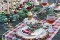 a bright Christmas tablescape with a plaid runner, evergreens, pinecones, gold candles and bright red and green blooms