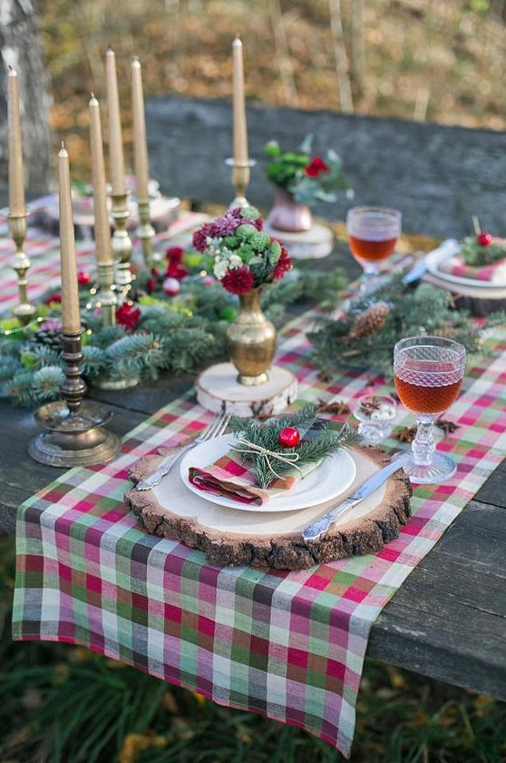 a bright Christmas tablescape with a plaid runner, evergreens, pinecones, gold candles and bright red and green blooms