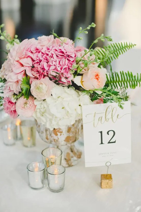 a bright wedding centerpiece of a gilded bowl, peachy ranunculus, pink and white hydrangeas, greenery and lots of candles around