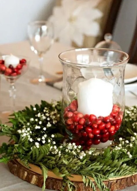 a chic rustic winter wedding centerpiece of a wood slice, greenery, baby’s breath and a glass with cranberries and a candle