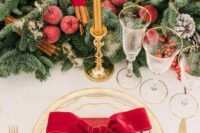 a luxurious Christmas wedding tablescape with an evergreen runner, sugared apples, berries and cinnamon sticks, gold rim plates and red napkins
