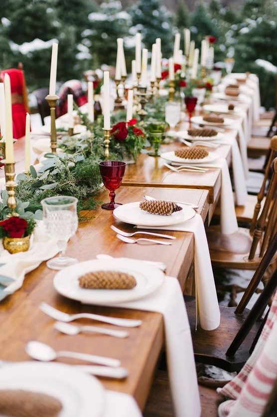 a refined Christmas tablescape with an evergreen runner, burgundy blooms and glasses, tall and thin candles and pinecones