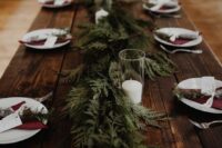 a rustic winter wedding tablescape with an uncovered table, burgundy napkins, an evergreen runner and candles in glasses