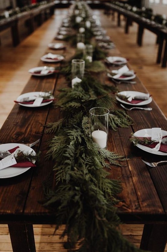 a rustic winter wedding tablescape with an uncovered table, burgundy napkins, an evergreen runner and candles in glasses