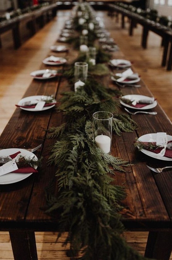 a very simple rustic tablescape with an uncovered table, a fern runner and evergreens, burgundy napkins