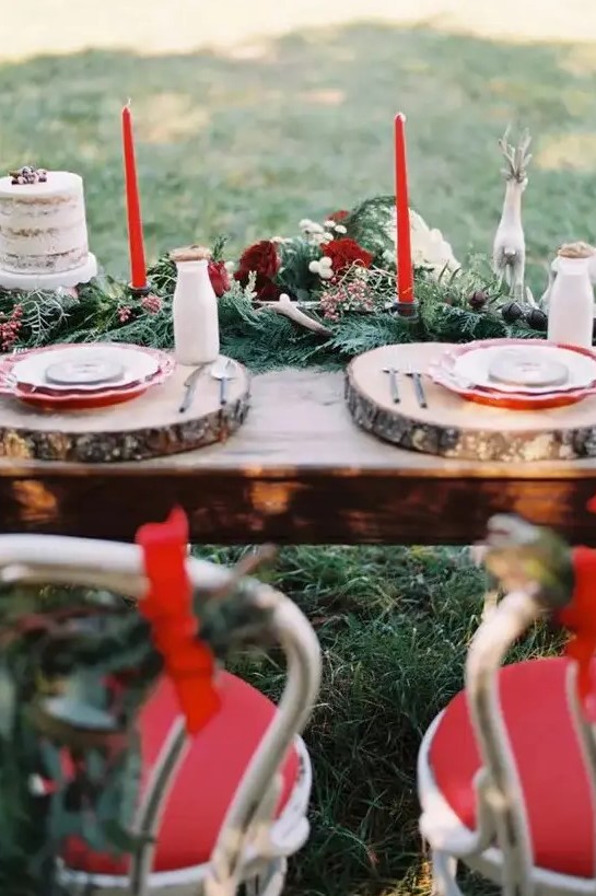 a winter wedding table setting with a greenery and fir runner, burgundy blooms and berries, red candles, wood slices