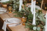 an evergreen and greenery table runner dotted with pinecones and with tall and thin candles is a beautiful and cozy idea for a winter wedding