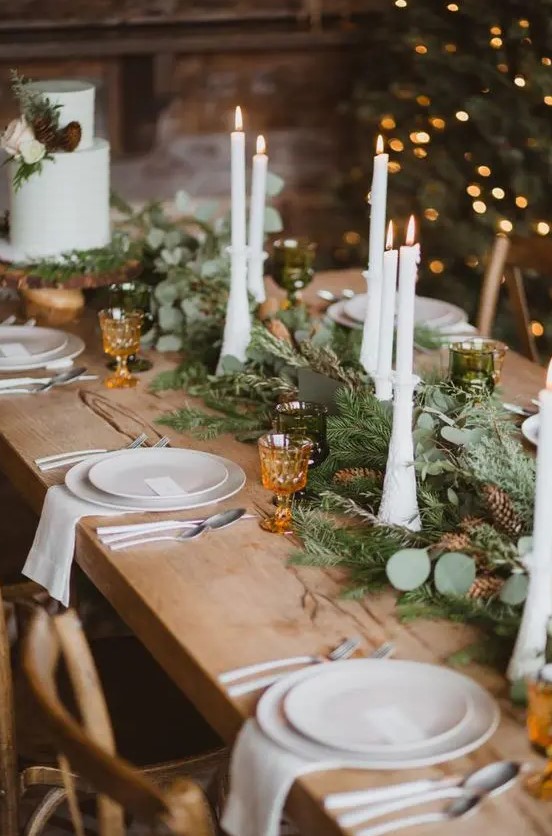 an evergreen and greenery table runner dotted with pinecones and with tall and thin candles is a beautiful and cozy idea for a winter wedding