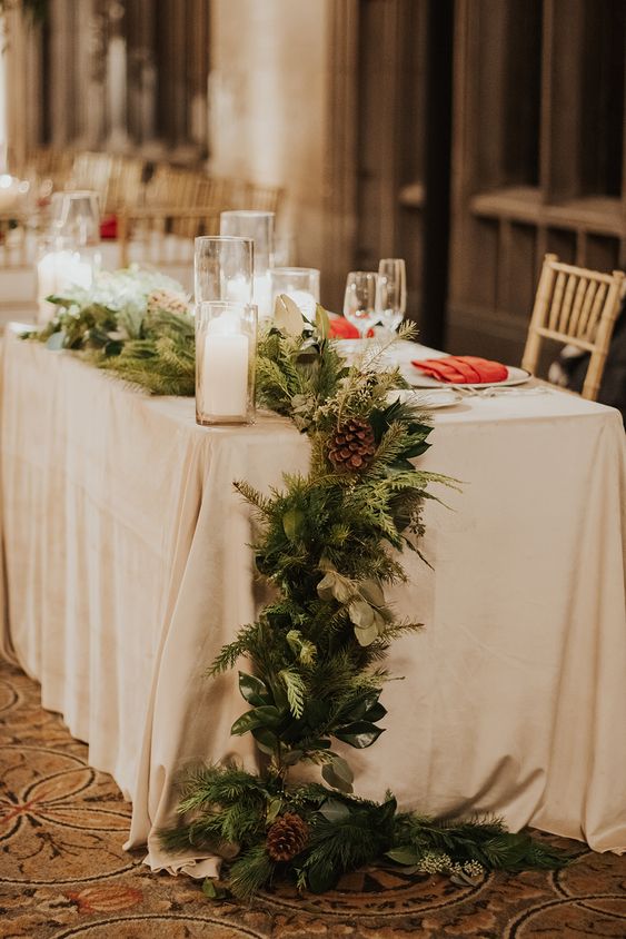 an evergreen and greenery table runner with pinecones and pillar candles is a classic decor idea for a winter wedding