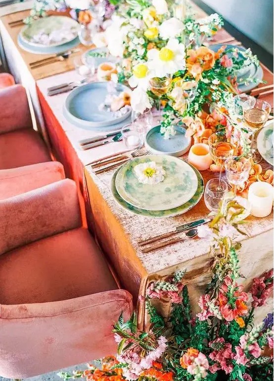 a bright wedding table setting with yellow, orange and white blooms, a peachy tablecloth and colorful plates, peachy candleholders and pink and orange blooms on the floor
