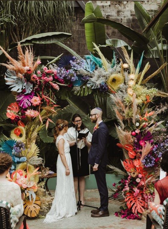 a bright and colorful wedding arch with bold blooms, pampas grass, tropical leaves and spray painted fronds is wow