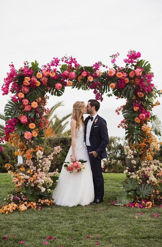 a colorful tropical wedding arch with hot pink, light pink, rust, yellow and orange blooms and monstera leaves is a gorgeous decor idea