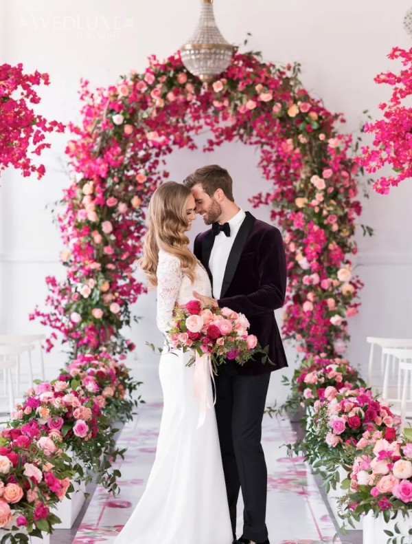 a bold wedding ceremony space with an arch covered with greenery, blush, hot pink and fuchsia blooms and planters with matching blooms
