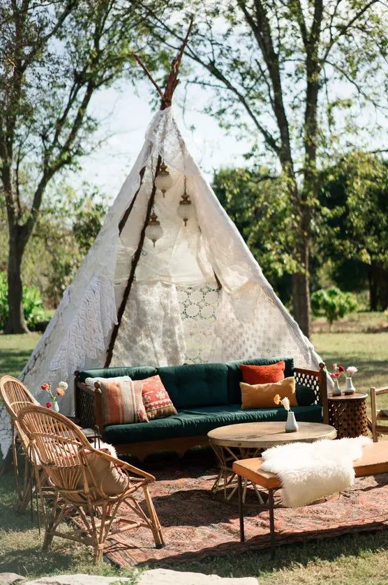 an outdoor boho wedding lounge with a green sofa, rattan chairs, a bench and a round coffee table, bright pillows and a doily teepee over the sofa
