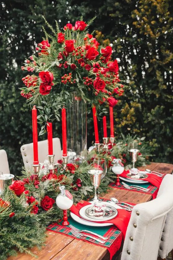a bold outdoor Christmassy wedding tablescape with an evergreen and red rose table runner, a tall matching centerpiece, red roses and red napkins